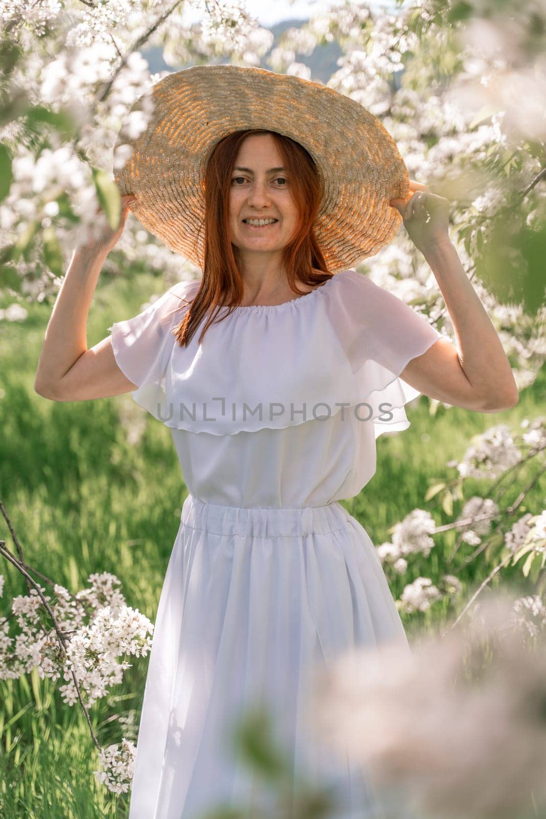 Woman cherry orchard. A happy woman in a long white long dress walks through the green spring blooming cherry garden. Happy cheerful princess bride. The fabric of the skirt flutters in the wind