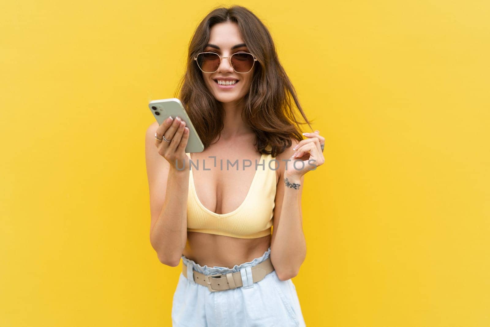 Young beautiful smiling woman with mobile phone. Carefree woman posing in the street near yellow wall. Positive model outdoors in sunglasses. Happy and excited