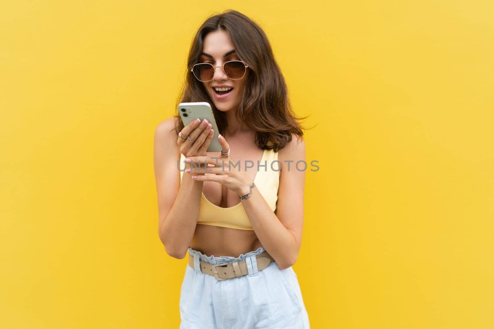 Young beautiful smiling woman with mobile phone. Carefree woman posing in the street near yellow wall. Positive model outdoors in sunglasses. Happy and excited