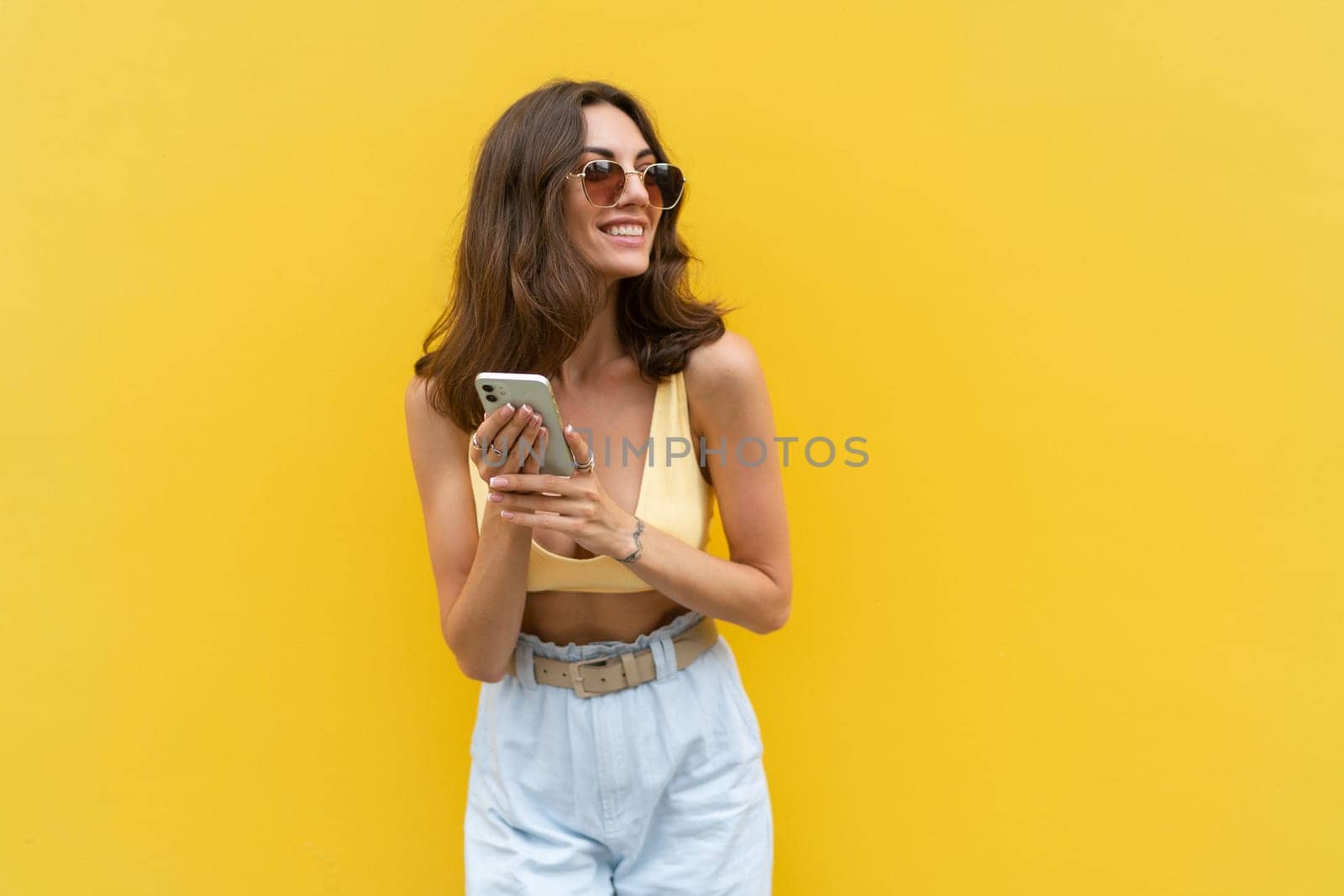 Young beautiful smiling woman with mobile phone. Carefree woman posing in the street near yellow wall. Positive model outdoors in sunglasses. Happy and excited