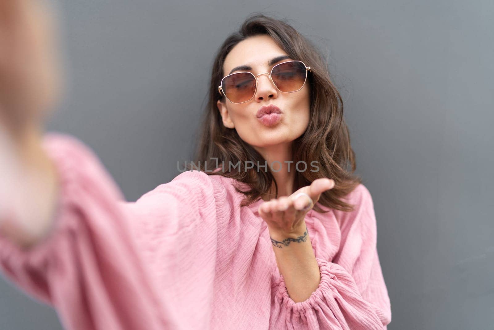 Young beautiful smiling cute romantic woman in trendy summer dress. Carefree woman posing in the street near grey wall. Positive model outdoors in sunglasses. Takes selfie.