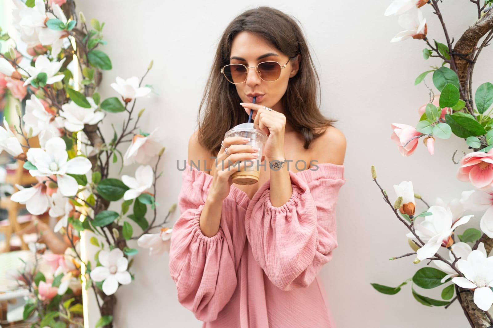 Young beautiful smiling cute romantic woman in trendy summer pink dress. Carefree woman posing in the street near white wall with flowers. Positive model outdoors in sunglasses. With cup of iced coffee latte. by kroshka_nastya
