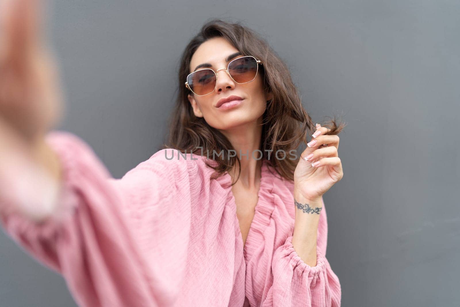Young beautiful smiling cute romantic woman in trendy summer dress. Carefree woman posing in the street near grey wall. Positive model outdoors in sunglasses. Takes selfie.