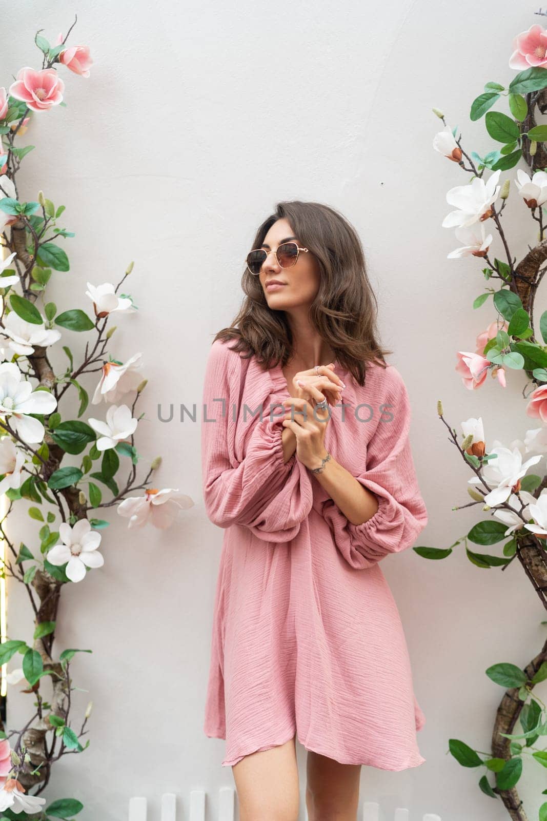 Young beautiful smiling cute romantic woman in trendy summer pink dress. Carefree woman posing in the street near white wall with flowers. Positive model outdoors in sunglasses.