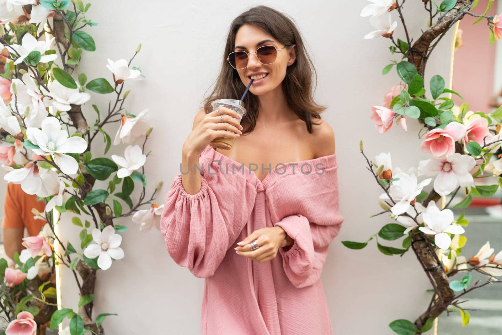 Young beautiful smiling cute romantic woman in trendy summer pink dress. Carefree woman posing in the street near white wall with flowers. Outdoors in sunglasses. With cup of iced coffee latte.