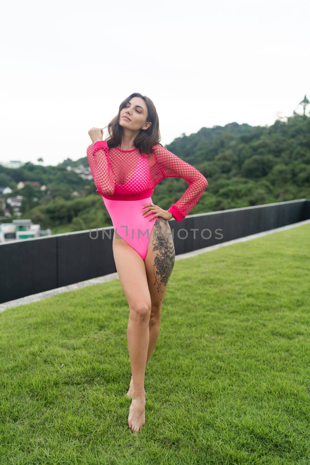 A slender young woman in a bright swimsuit on the grass by the pool, a large tattoo on her thigh. Slim athletic girl posing, tender romantic sexy