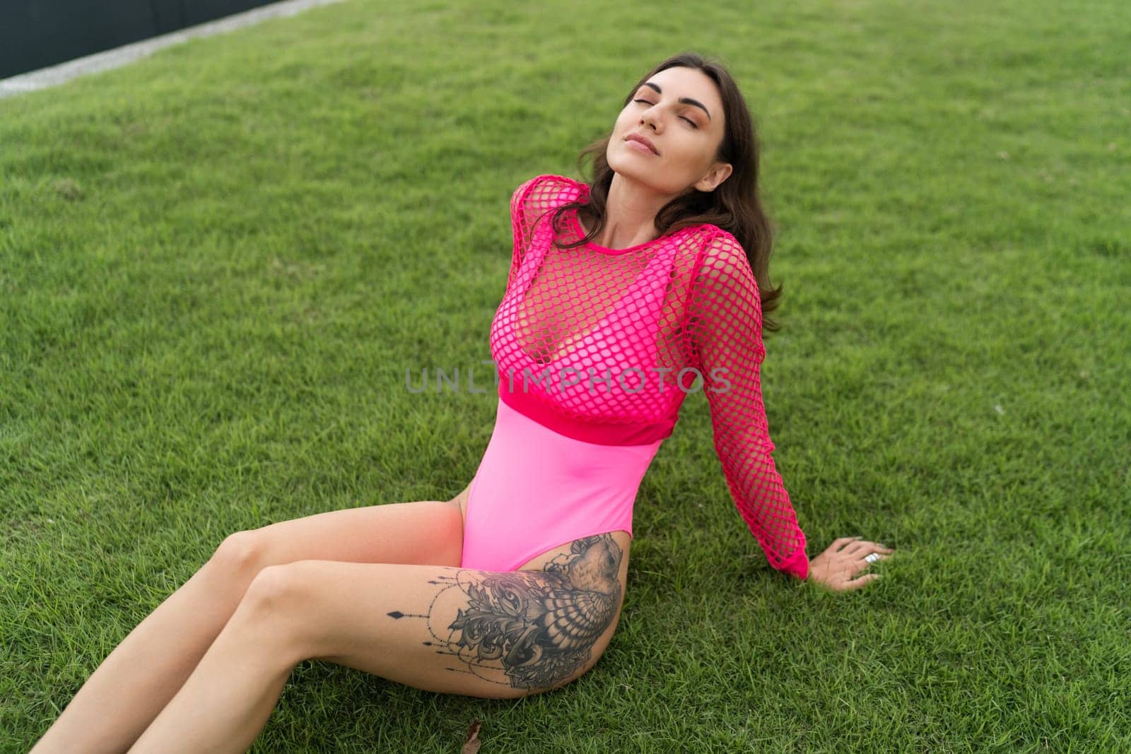 A slender young woman in a bright swimsuit on the grass by the pool, a large tattoo on her thigh. Slim athletic girl posing, tender romantic sexy