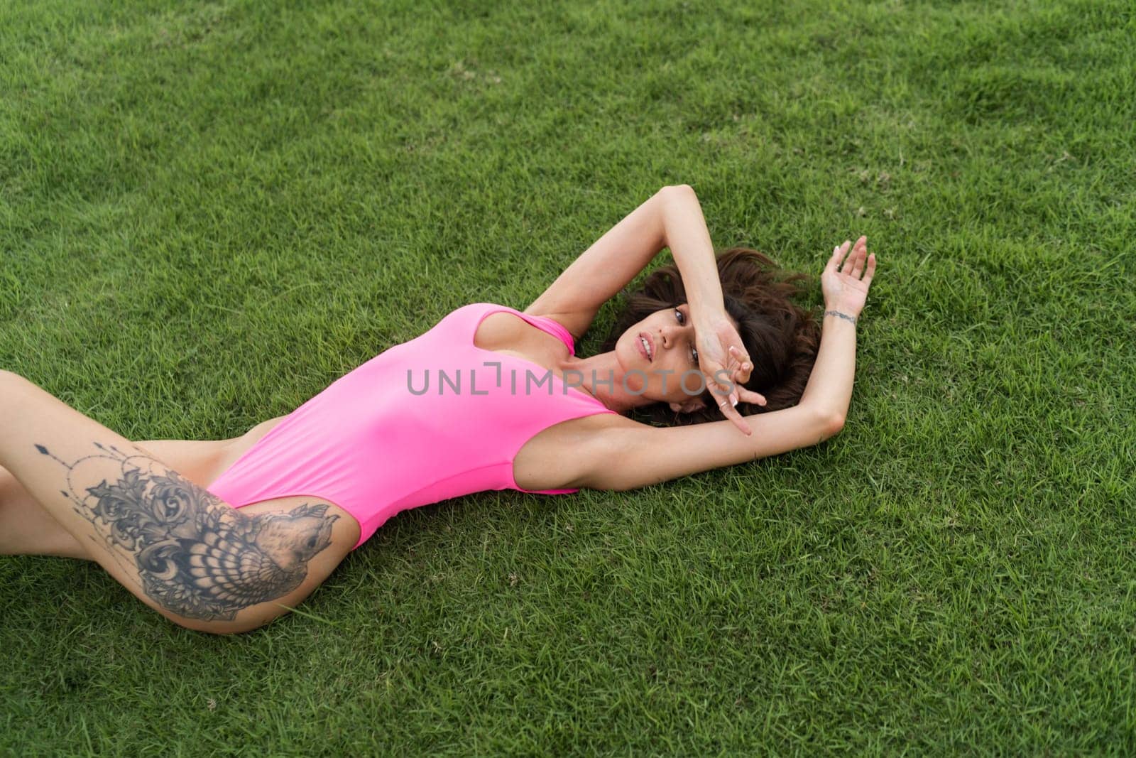 A slender young woman in a bright swimsuit on the grass by the pool, a large tattoo on her thigh. Slim athletic girl posing, tender romantic sexy