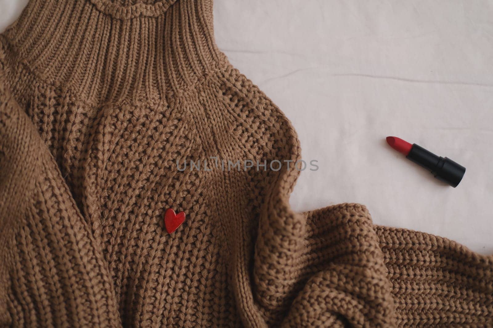 Overhead view of woman's casual outfit with warm knitted sweater, red lipstick and earrings on beige background in bed, Flat lay, top view by paralisart