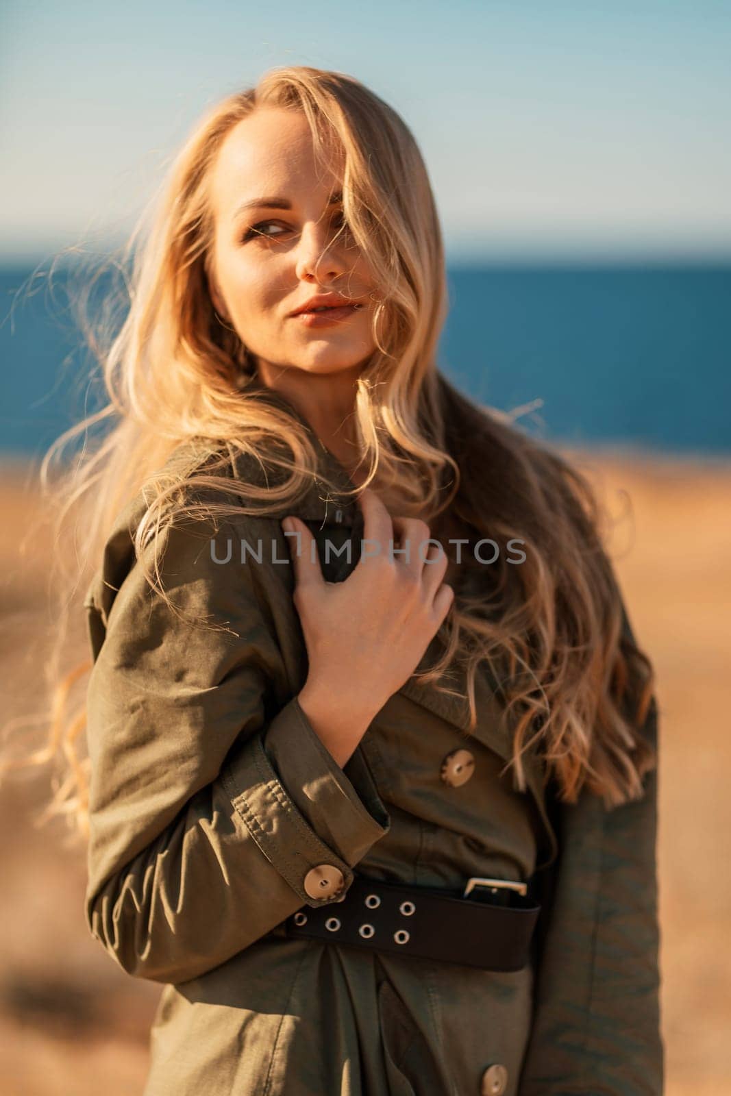 Portrait blonde sea cape. Calm young blonde in a khaki raincoat stands on the seashore.