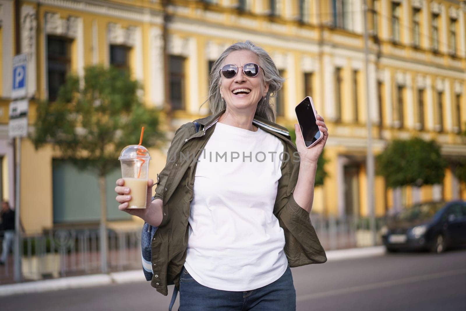A Mature European Woman Enjoys Life Walking in The City. Biohacking Concept. Mature gray-haired modern european woman walks around the city in sunglasses, holding a mobile phone and a drink with a straw. High quality photo.