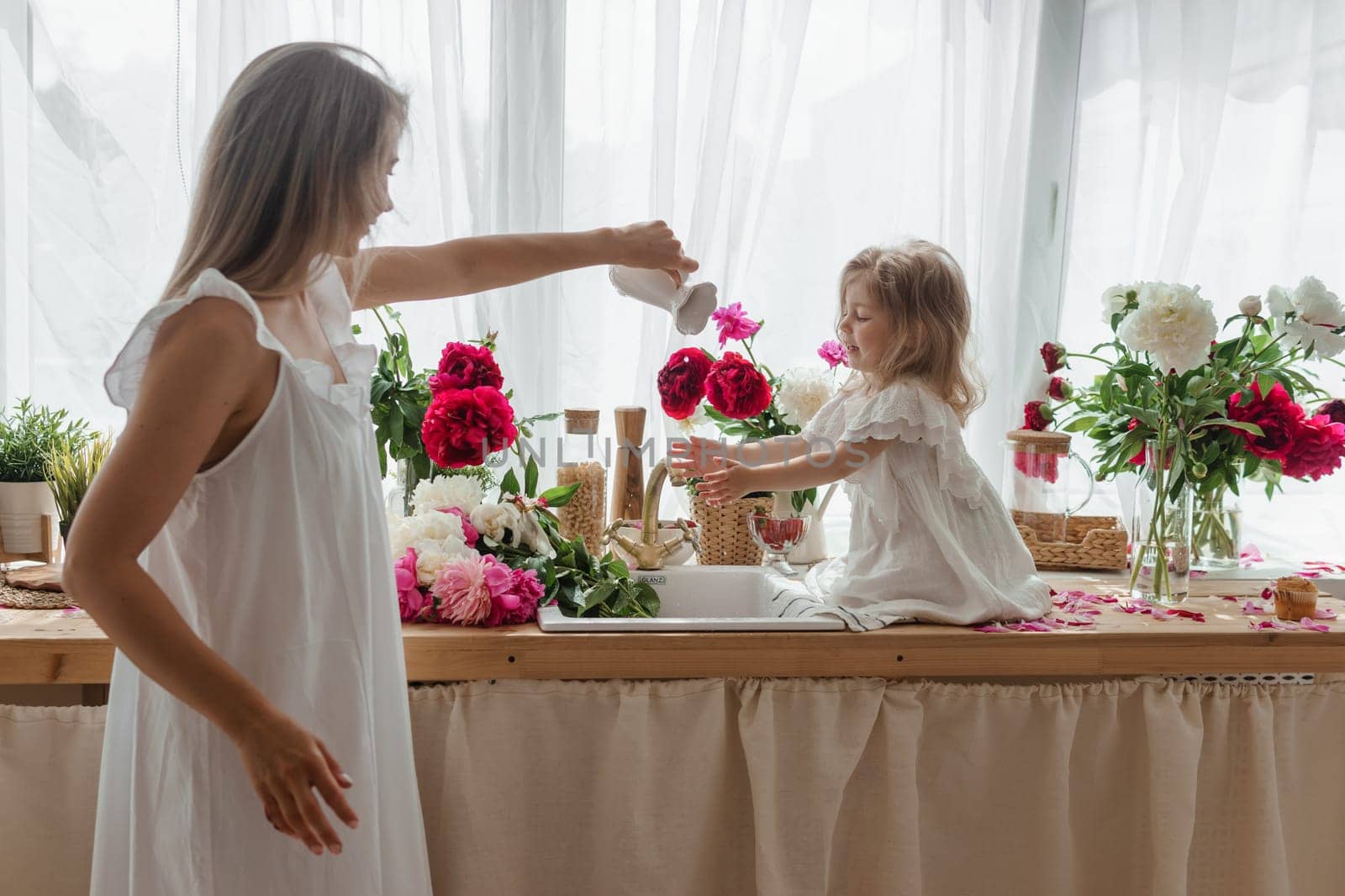 A little blonde girl with her mom on a kitchen countertop decorated with peonies. The concept of the relationship between mother and daughter. Spring atmosphere.