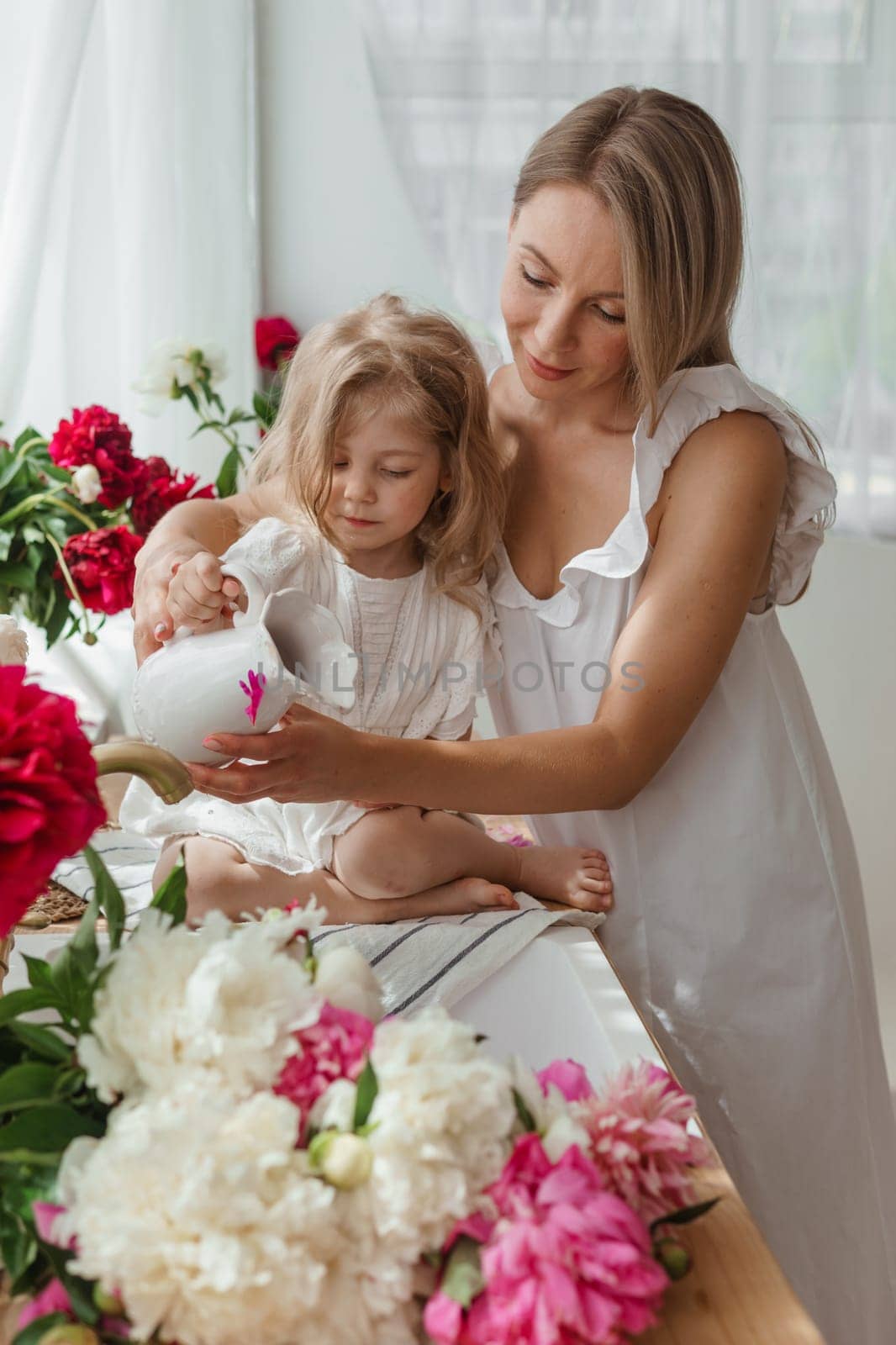 A little blonde girl with her mom on a kitchen countertop decorated with peonies. The concept of the relationship between mother and daughter. Spring atmosphere.