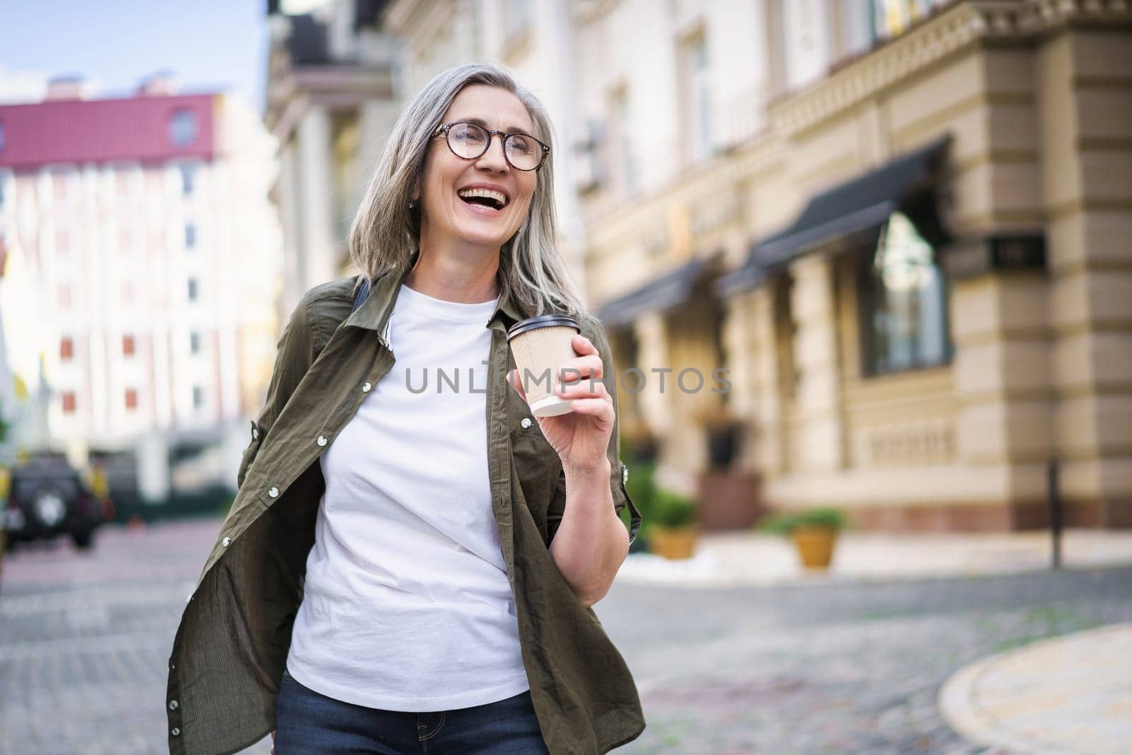 Stylish senior woman with grey hair holding coffee cup while enjoying vibrant atmosphere of European city. Colorful cityscape and architectural landmarks create timeless and classic background that highlights beauty, grace, and dignity of aging.
