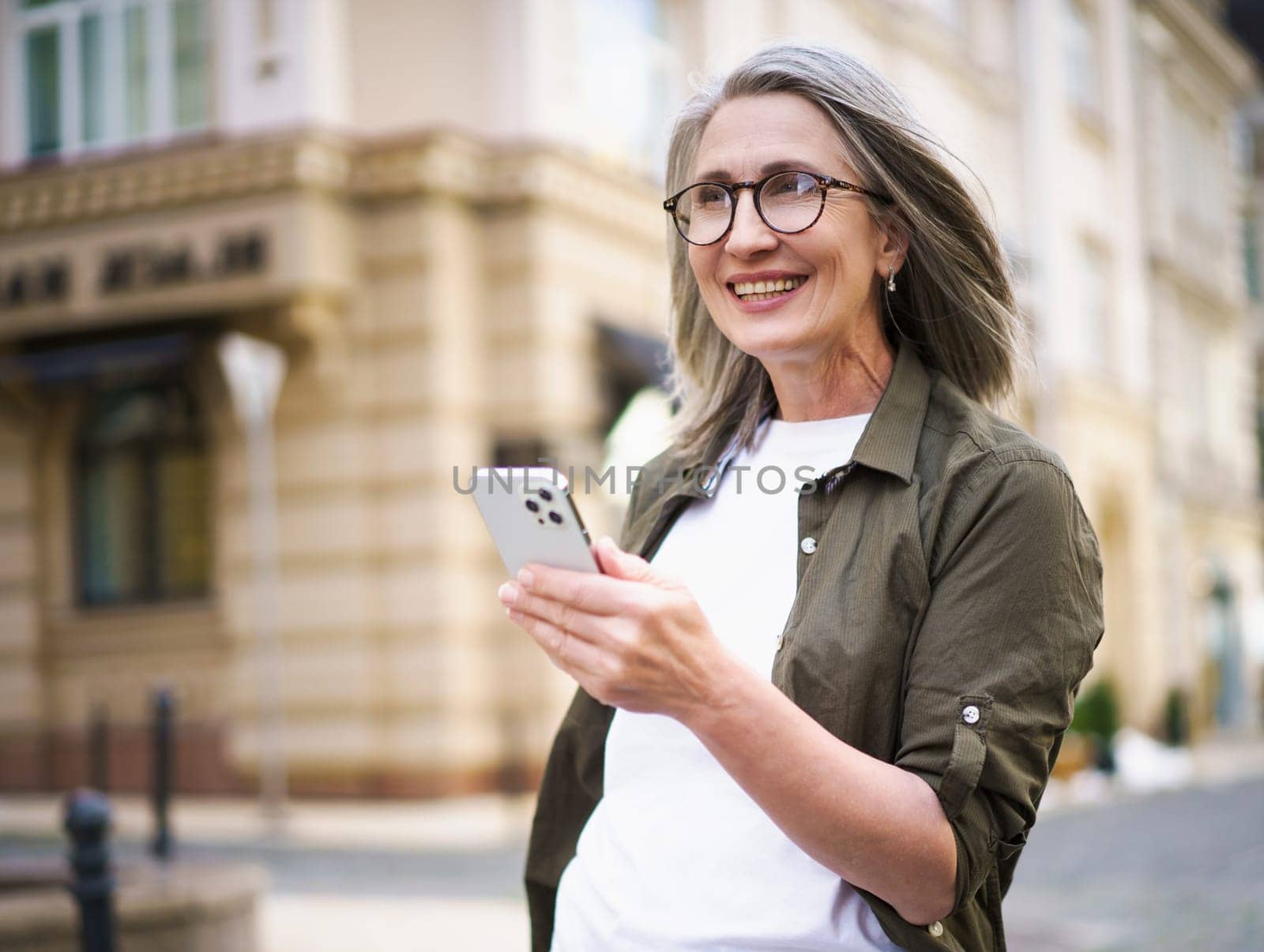 Joyful senior woman with grey hair and mobile phone in hand, enjoying her time in beautiful European city. Image showcases active and modern urban lifestyle of the elderly generation. High quality photo
