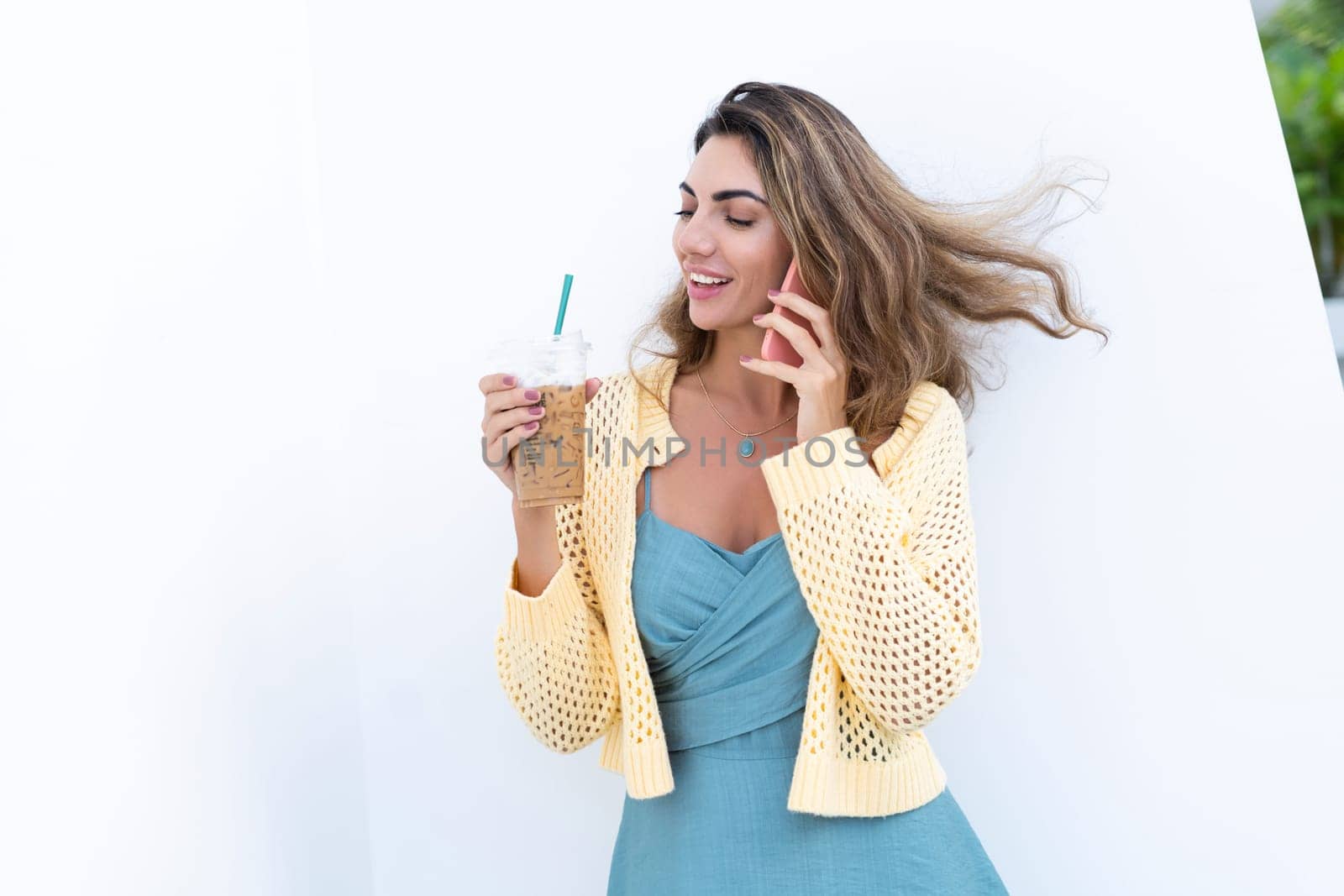 Portrait of beautiful woman in green summer dress on white background natural daylight, holding iced coffee cappuccino talking on phone smiling