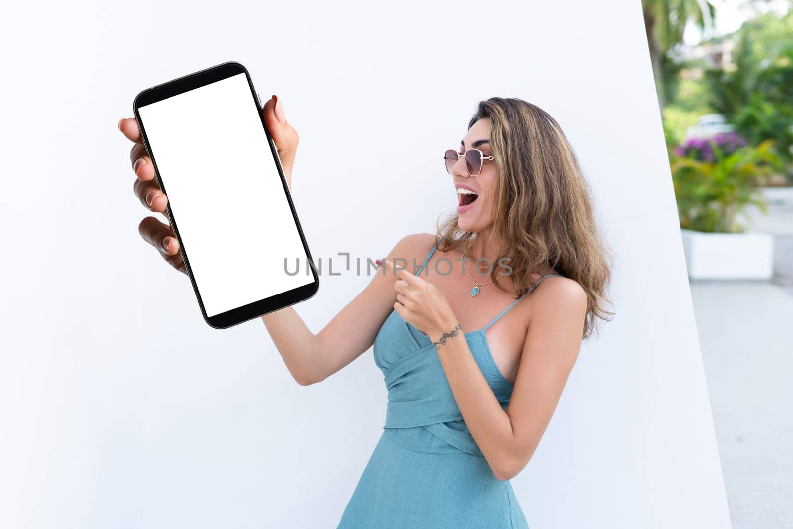 Portrait of beautiful woman in green summer dress on white background natural daylight, pointing on empty white big screen of smartphone, space for advertising by kroshka_nastya