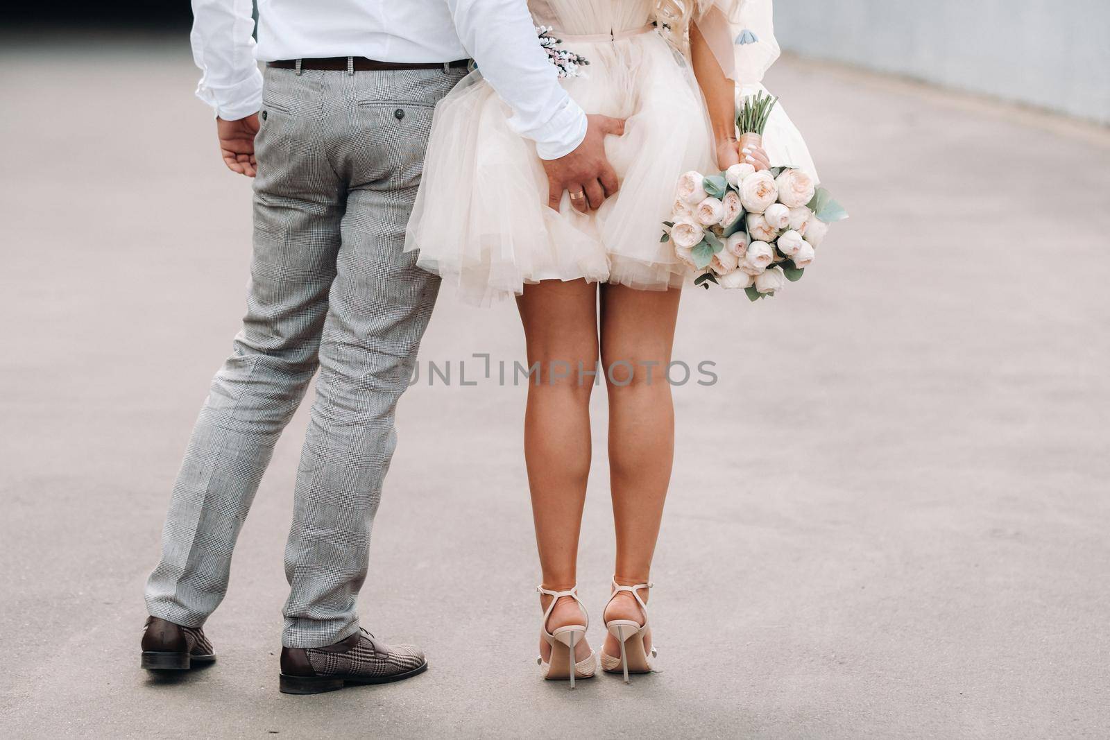 The groom puts his hand behind his bride. The bride in a short dress is holding a bouquet in her hands from behind.