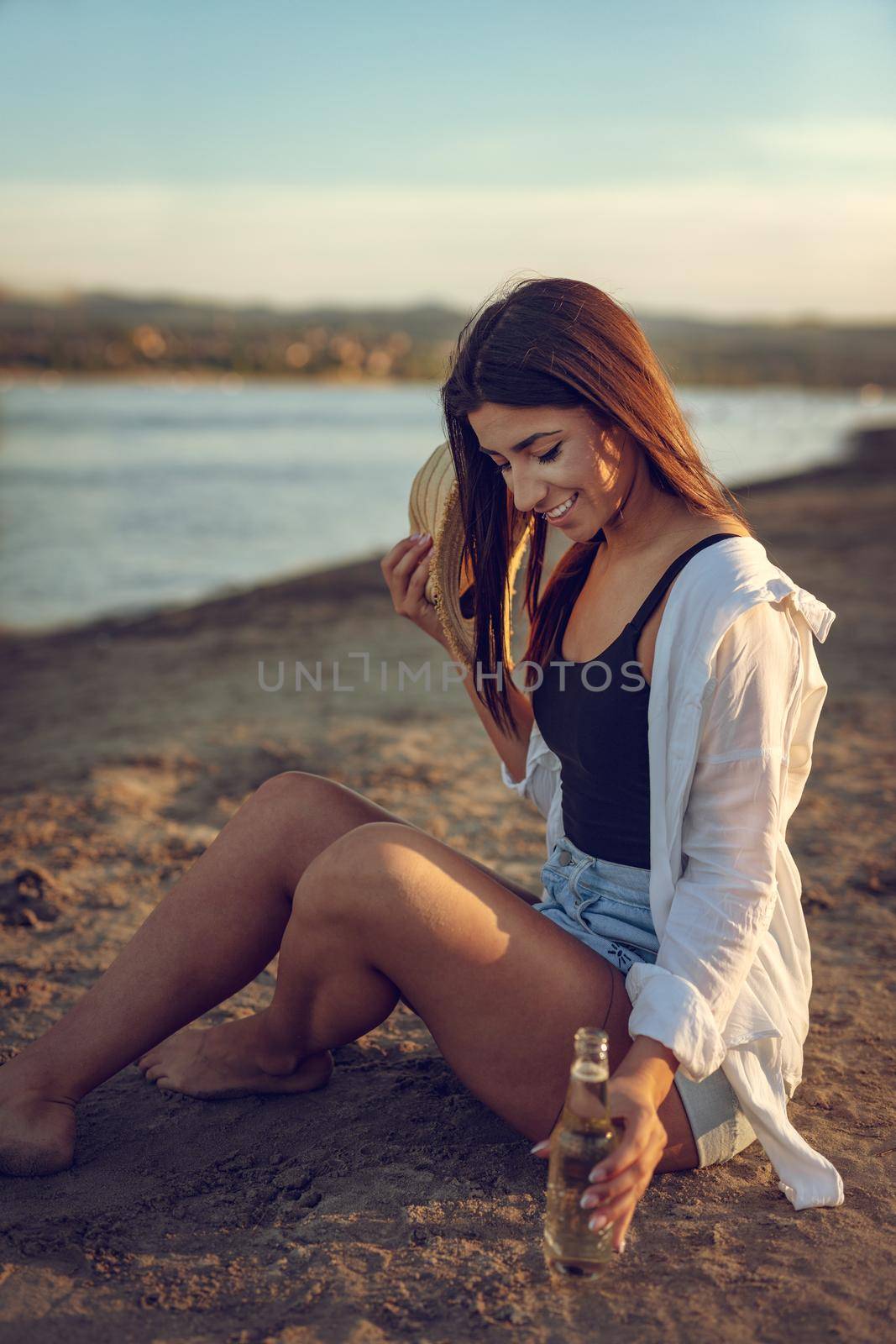 Young woman in straw hat relaxing at sunset time on the river bank. She is sitting on the river send and holding bottle of beer. 