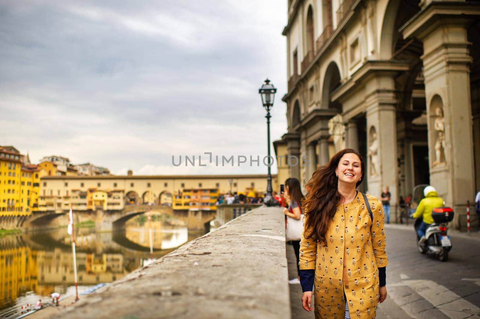 A tourist girl with an orange coat happily walks on the embankment in Florence, Italy by Lobachad