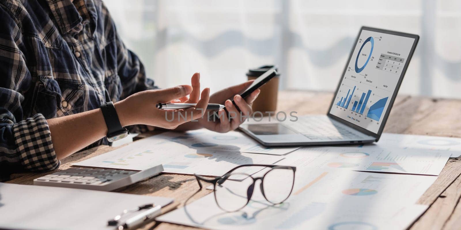 Young accountant hand holding smartphone to call marketing consultants and using laptop computer to analyze sales growth in the global workplace market. accounting concept by wichayada