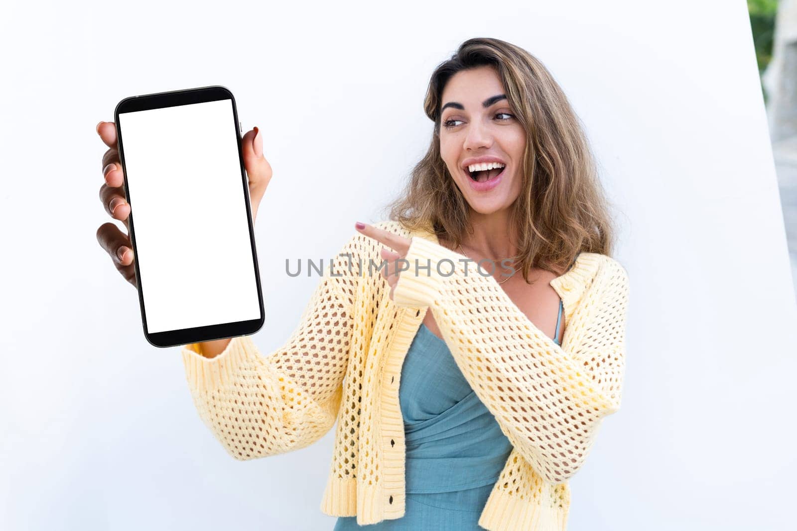 Portrait of beautiful woman in green summer dress on white background natural daylight, pointing on empty white big screen of smartphone, space for advertising