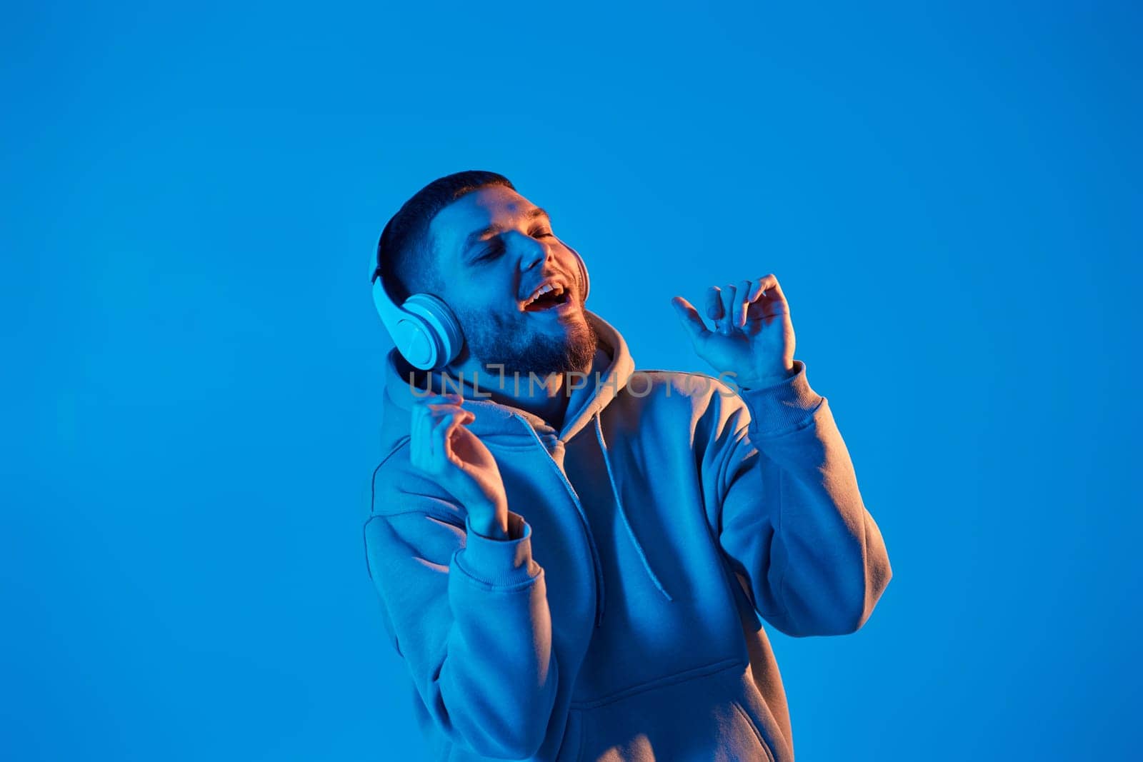 handsome bearded man with headphones in sweatshirt enjoying favorite music on blue neon background. Neon lighting