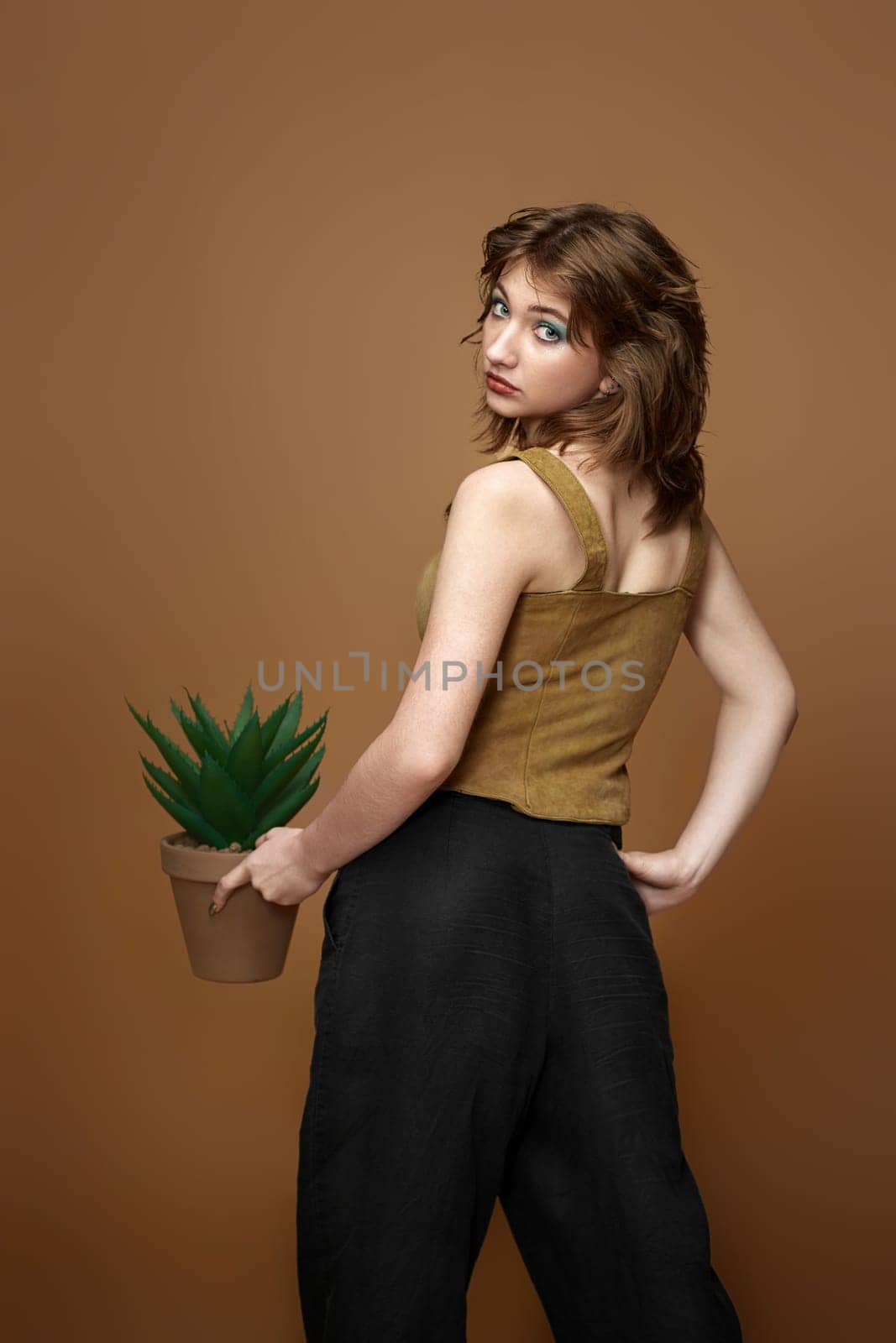 young woman with stylish hairstyle posing with plant in pot on beige background.