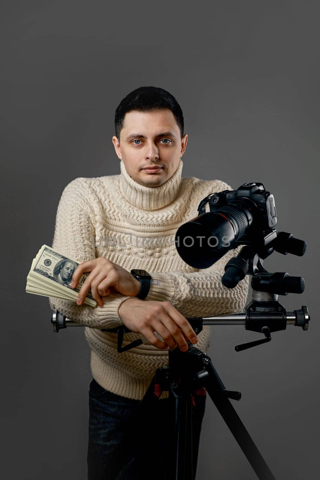 Professional photographer with digital camera on tripod holding hundred dollar bills on gray background
