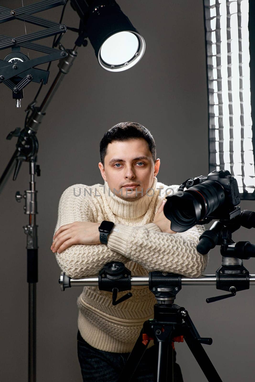 Professional photographer with digital camera on tripod looking at the camera on gray background with lighting equipment