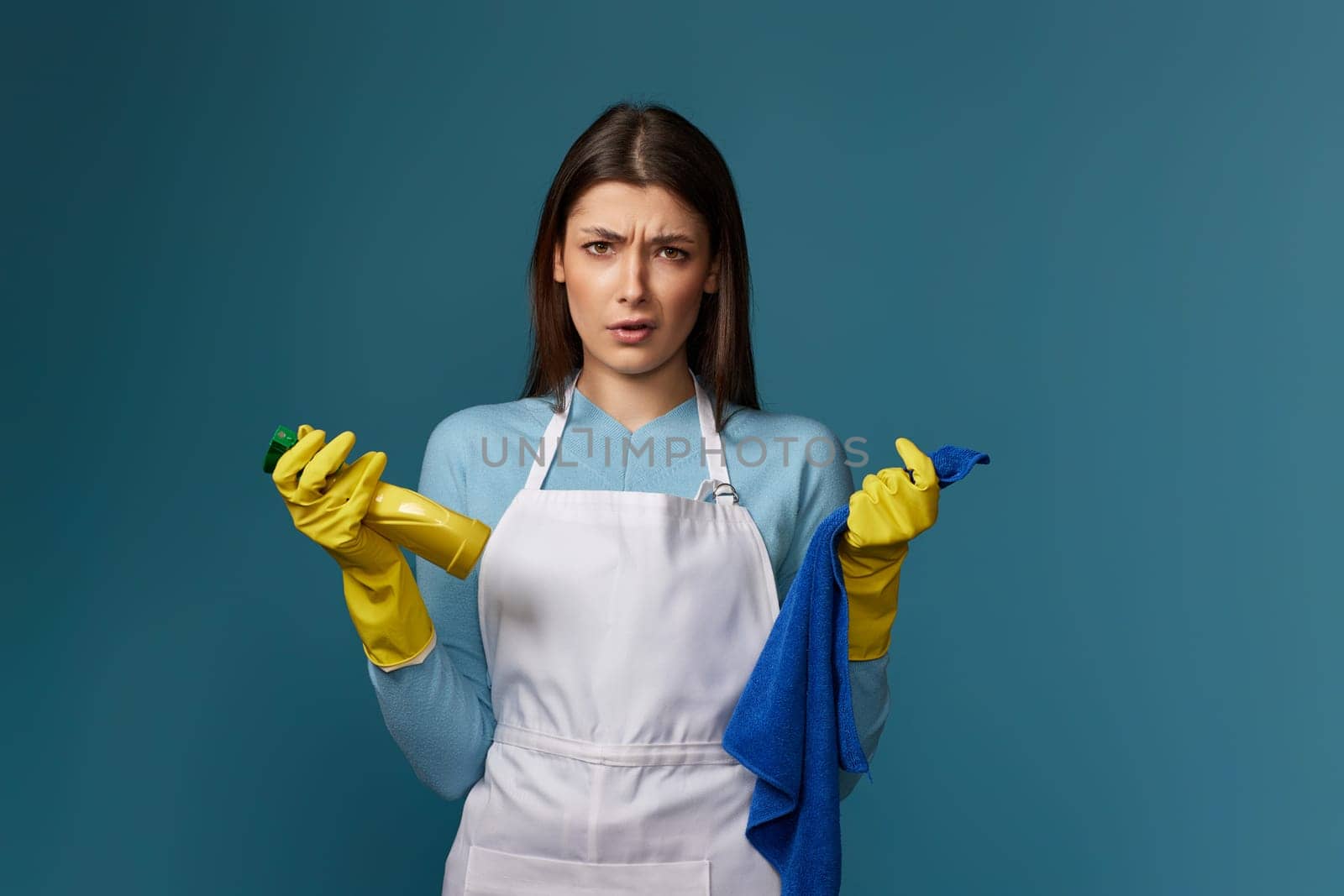 skeptic and nervous woman in gloves and cleaner apron with cleaning rag and detergent sprayer on blue background.