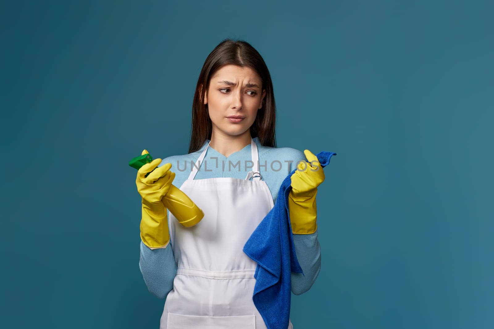 skeptic and nervous, frowning young woman in cleaner apron with cleaning rag and detergent sprayer on blue background.