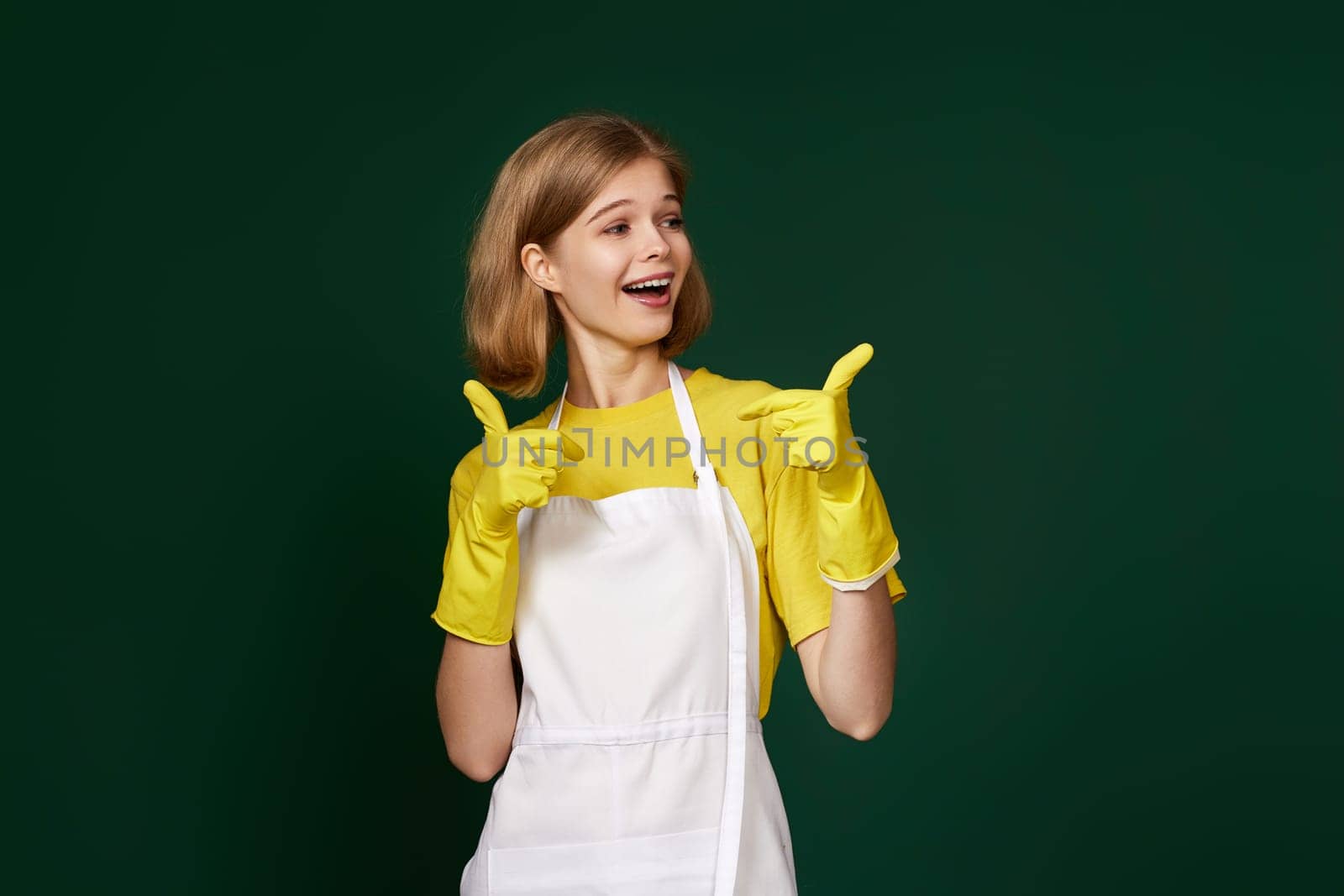 beautiful blonde woman in yellow rubber gloves and cleaner apron showing ok sign on green background.