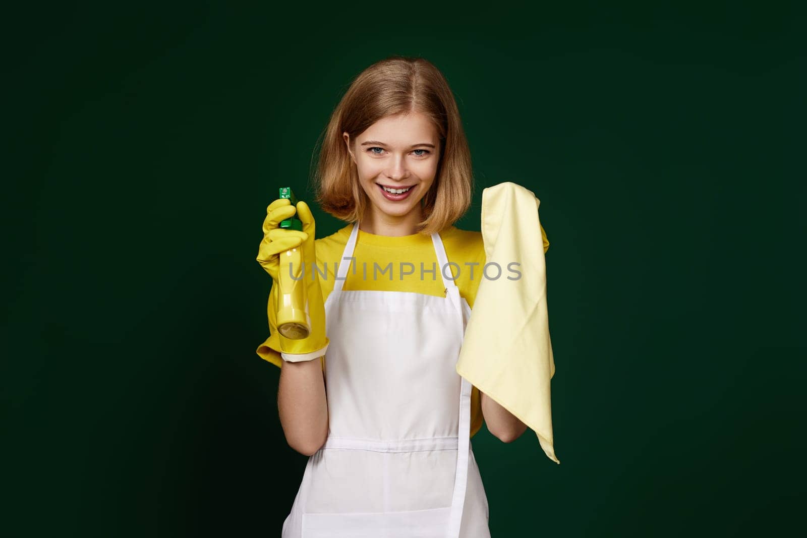happy blonde woman in yellow rubber gloves and cleaner apron with cleaning rag and detergent sprayer on dark green background.