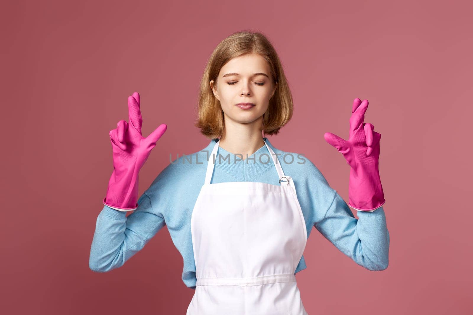 beautiful blonde caucasian girl in rubber gloves with finger crossed gesture on pink background.