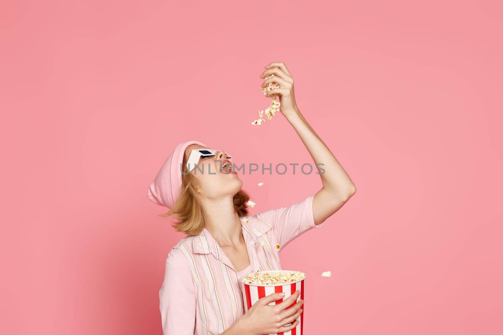 pleased blonde woman in t-shirt and hat eating popcorn on pink background.