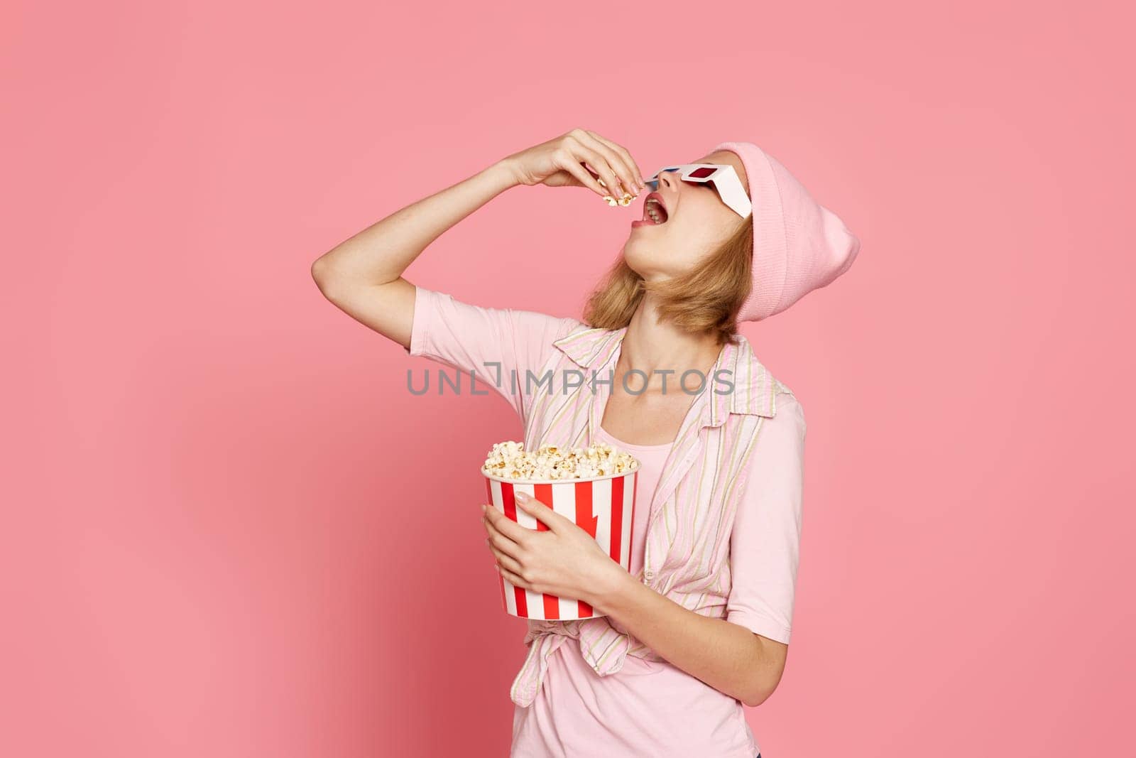 happy blonde woman in 3d glasses eating popcorn on pink background.