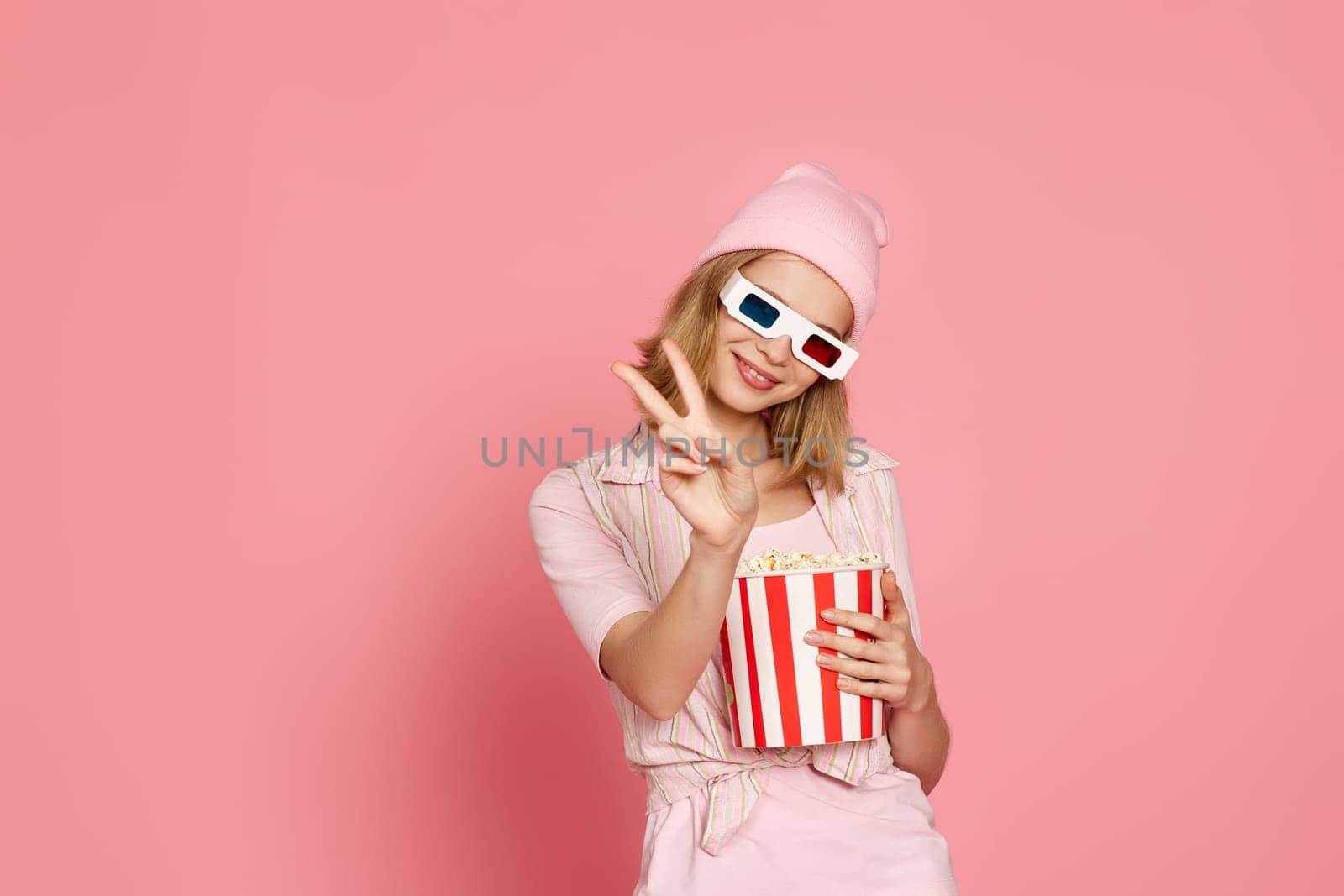 attractive blonde woman in 3d glasses holding bucket of popcorn and showing victory gesture on pastel pink background