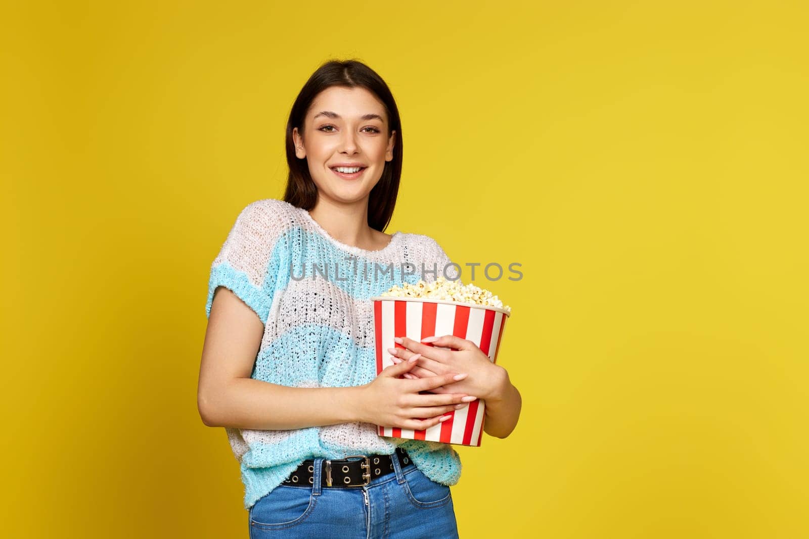 smiling brunette girl in blue pullover holding bucket of popcorn on yellow background