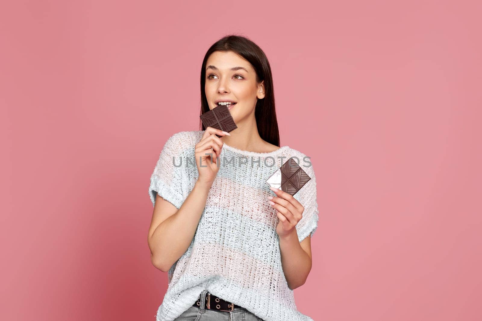 charming happy woman in sweater biting sweet chocolate bar and looking at the side isolated on pastel pink background