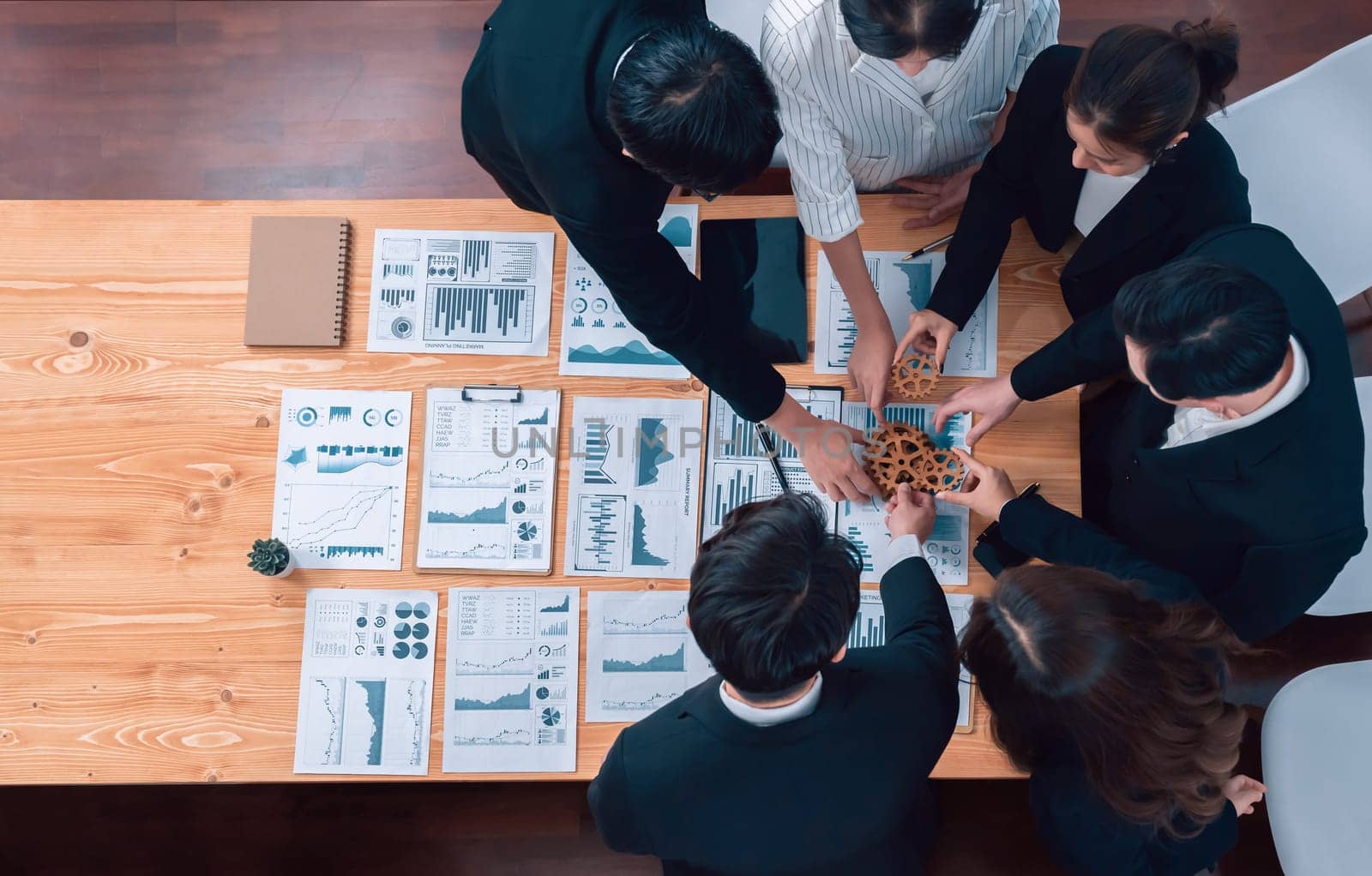 Top view hand holding gear by businesspeople wearing suit for harmony synergy in office workplace concept. Group of people hand making chain of gears into collective form with dashboard report papers.