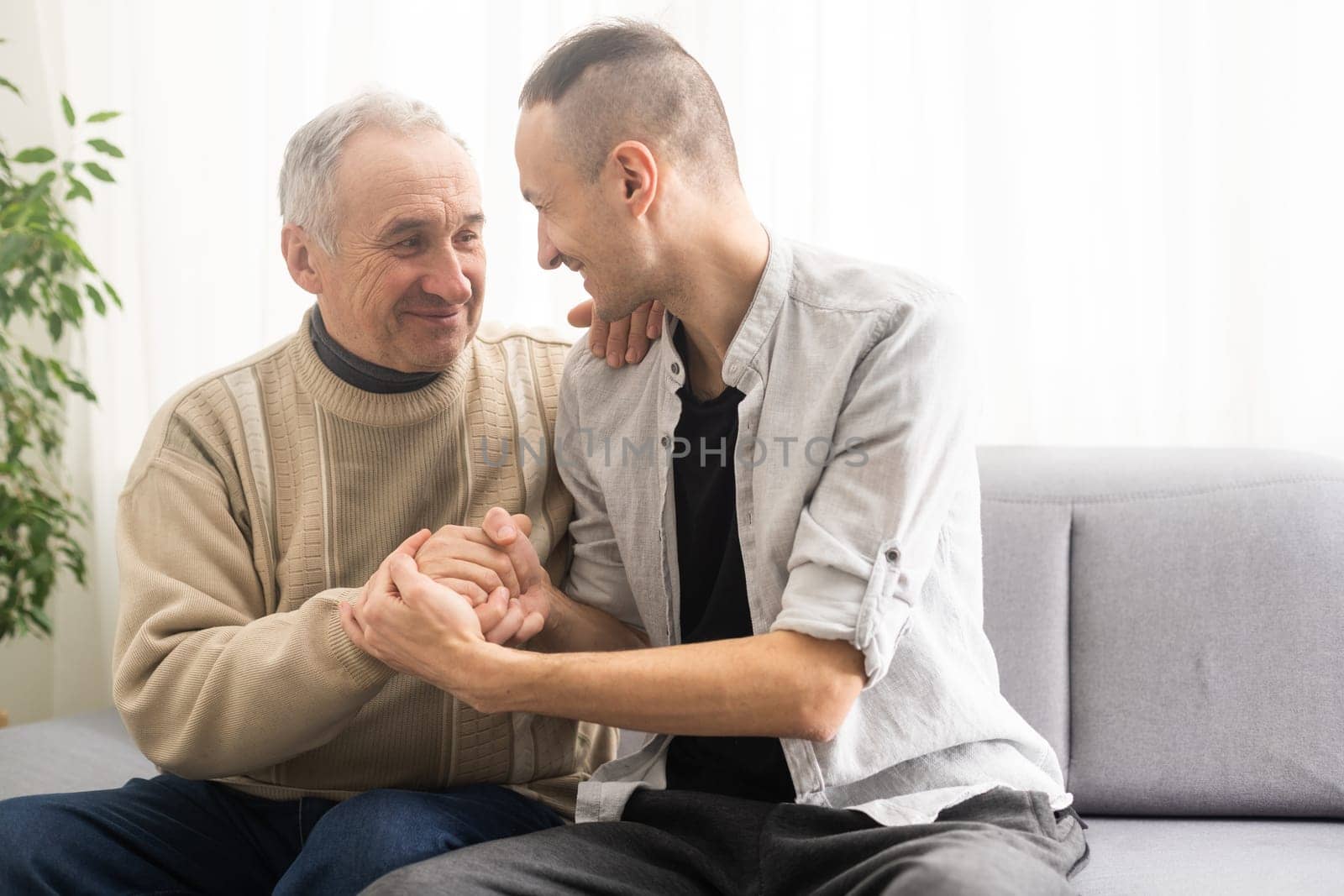 Happy two generations male family old senior mature father and smiling young adult grown son enjoying talking chatting bonding relaxing having friendly positive conversation sit on sofa at home