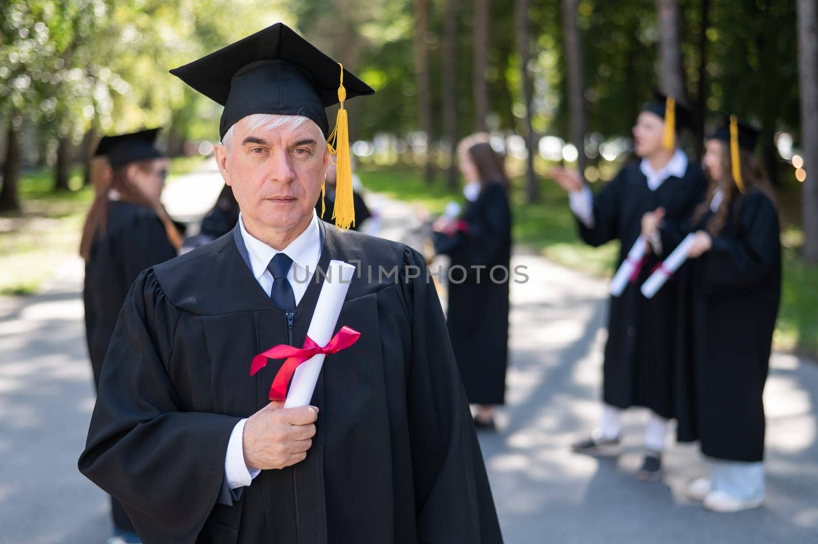 A group of graduates in robes outdoors. An elderly student rejoices at receiving a diploma. by mrwed54