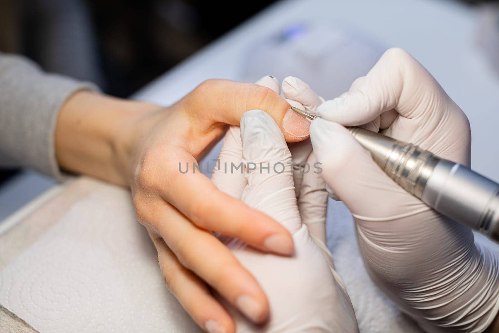 A beautician removes cuticles from a thumb nail. A beauty salon customer has a manicure done. The nail milling machine effectively prepares the nail plate. The beautician wears disposable gloves.