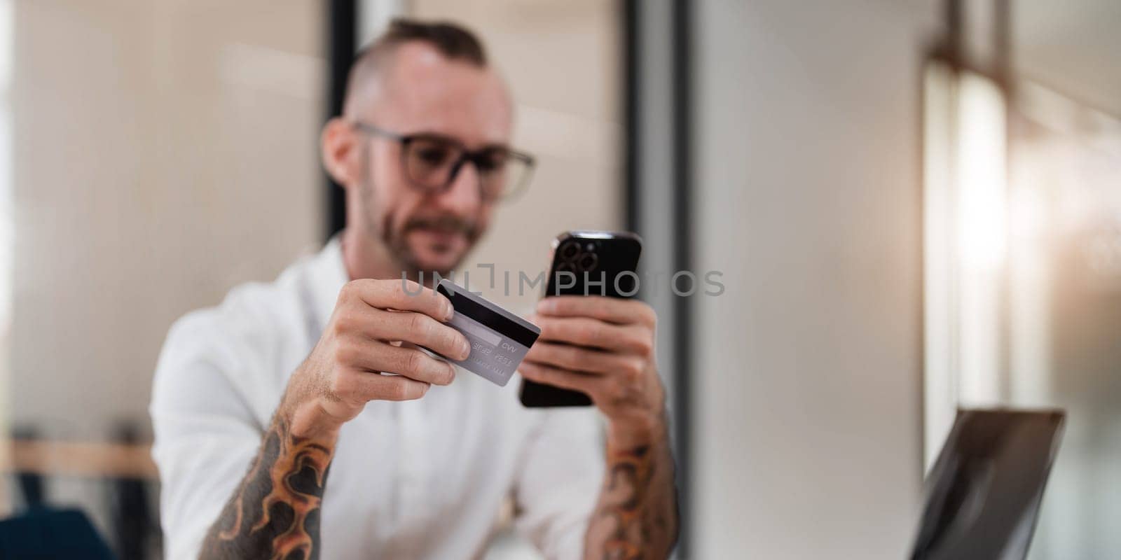 A man reading information from the back of the credit card and typing it on the smartphone to do an online purchase at home. A guy doing an online payment on a cellphone at table in office..