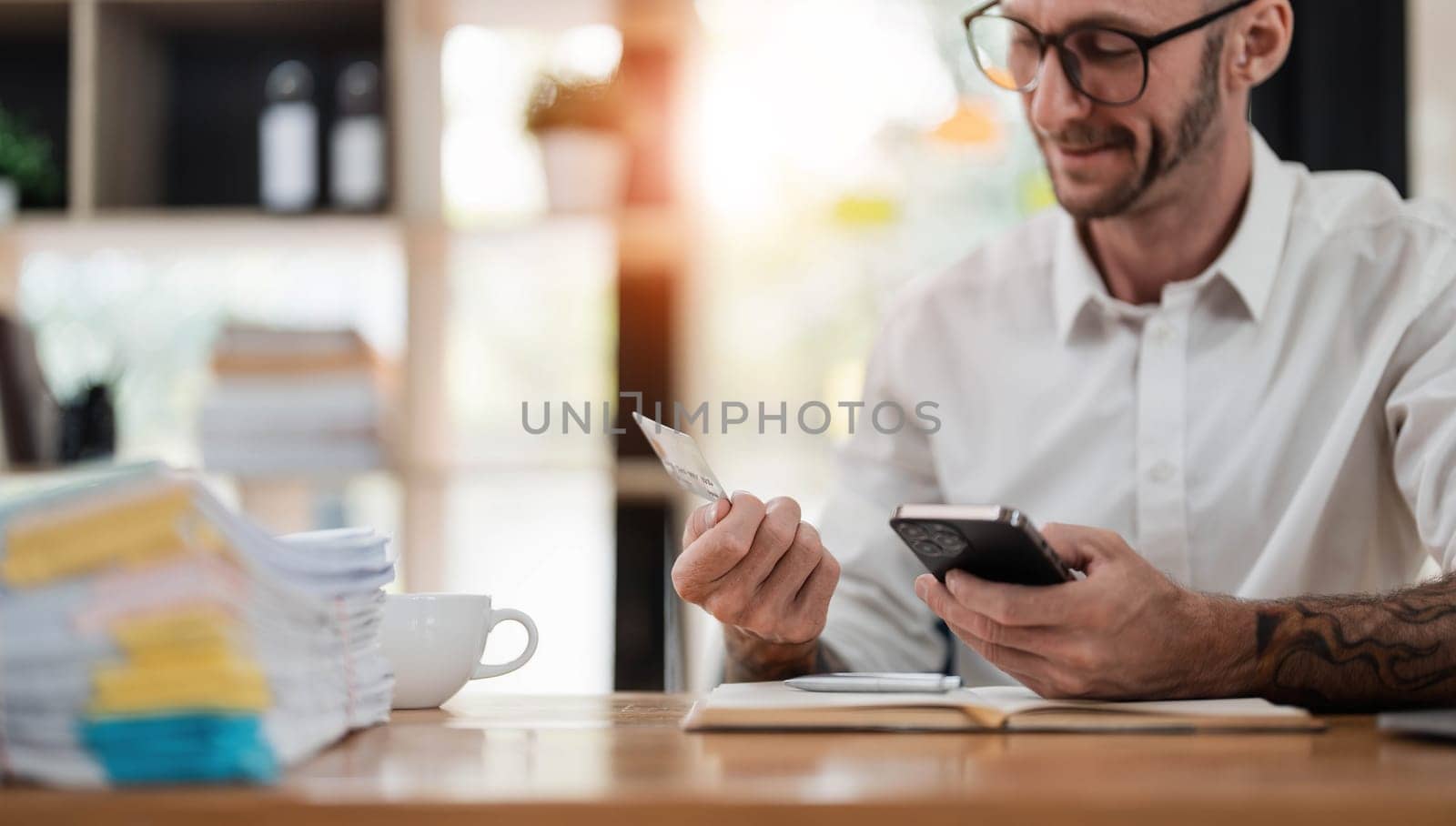 A man reading information from the back of the credit card and typing it on the smartphone to do an online purchase at home. A guy doing an online payment on a cellphone at table in office..