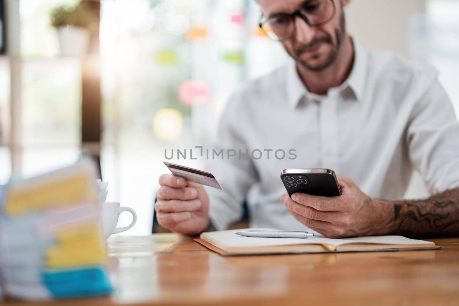A man reading information from the back of the credit card and typing it on the smartphone to do an online purchase at home. A guy doing an online payment on a cellphone at table in office by wichayada