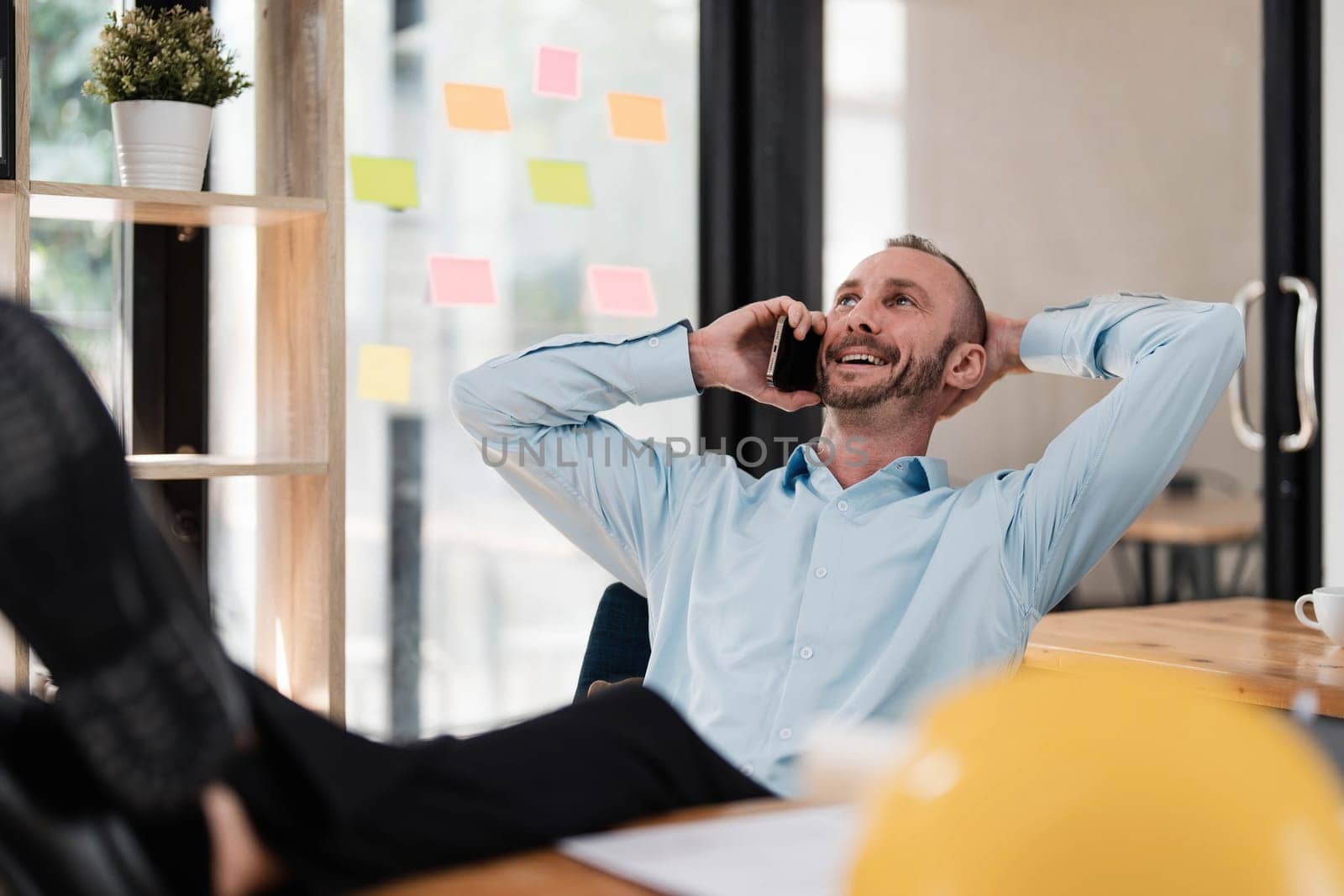 Handsome and happy Caucasian male engineer putting his legs on table and hand behind head while talking on the phone with client and chilling in the office. by wichayada