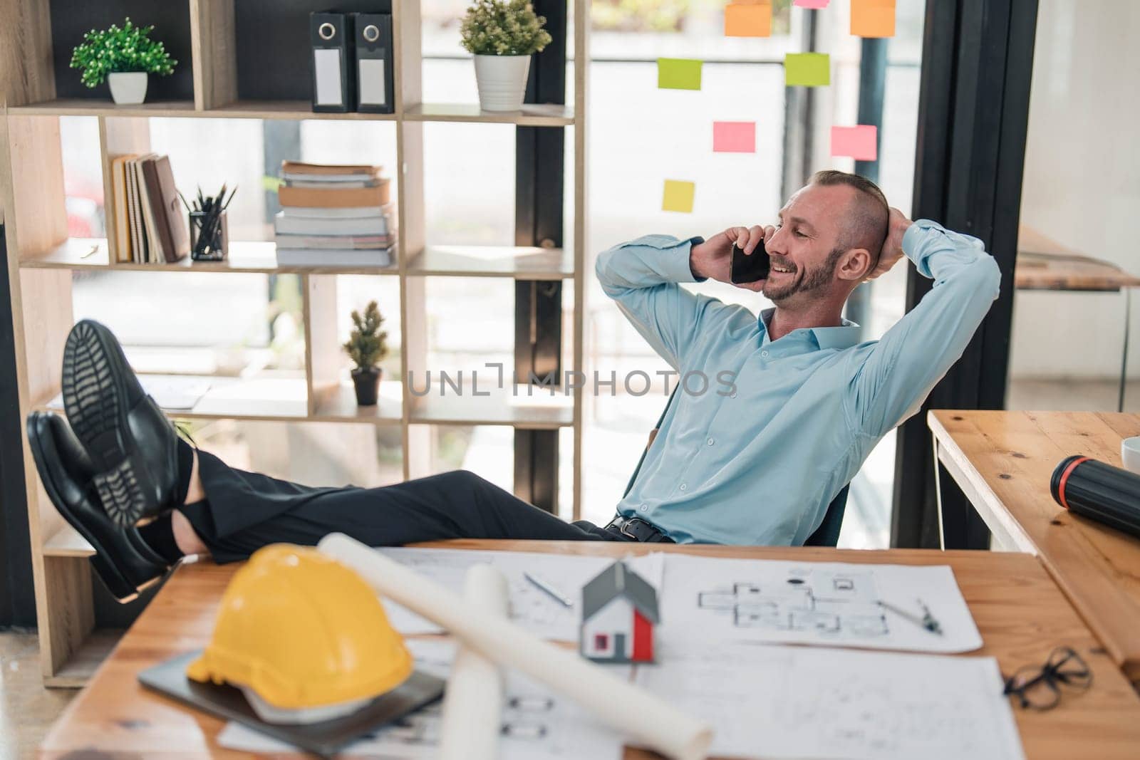 Handsome and happy Caucasian male engineer putting his legs on table and hand behind head while talking on the phone with client and chilling in the office. by wichayada