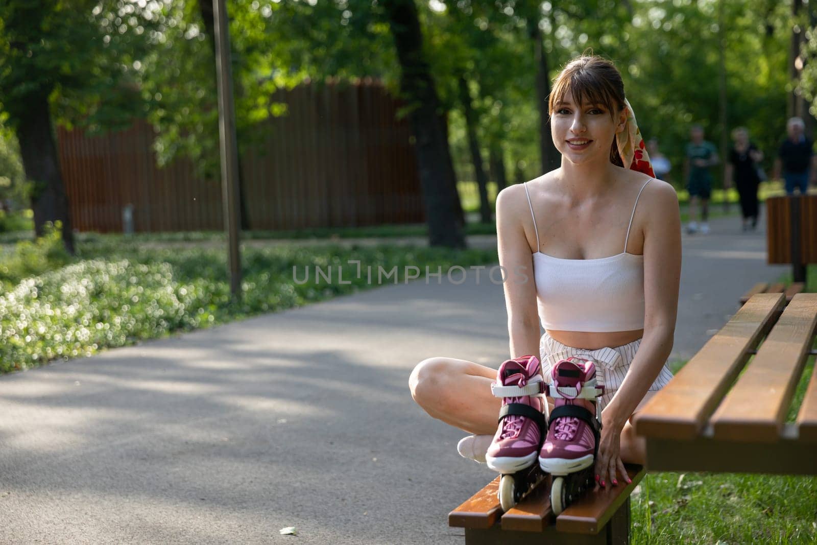 A smiling young woman sits on a bench with her legs crossed. The girl holds rollerblades standing on the bench in front of her. In the background you can see a green park, trees, lawns and an asphalt alley.